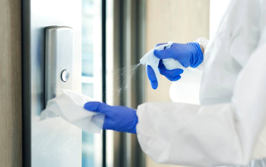 A person in latex gloves and protective gown sprays disinfectant on a cloth to clean an elevator button for coronavirus.