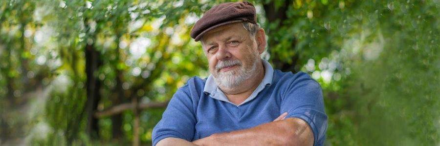 A solemn elderly gentleman wearing a beret stands with folded arms stands before a grouping of trees and shrubs in an outdoor setting.