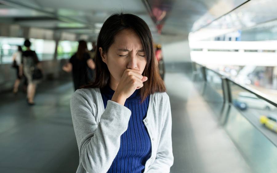 A woman coughs into her hand inside a building during flu season.