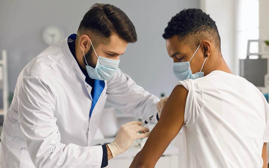 A young African-American man gets vaccinated by health care provider wearing a mask.