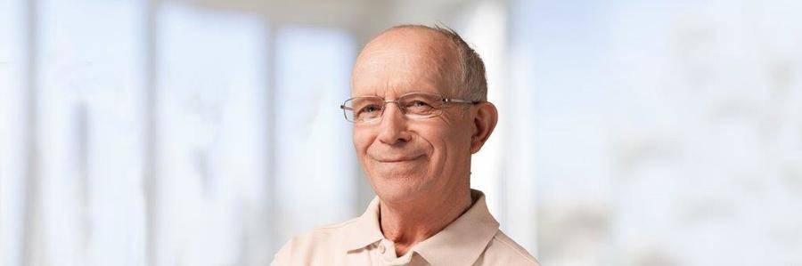 A smiling elderly gentleman wearing spectacles relaxes before a brightly lit bank of windows.