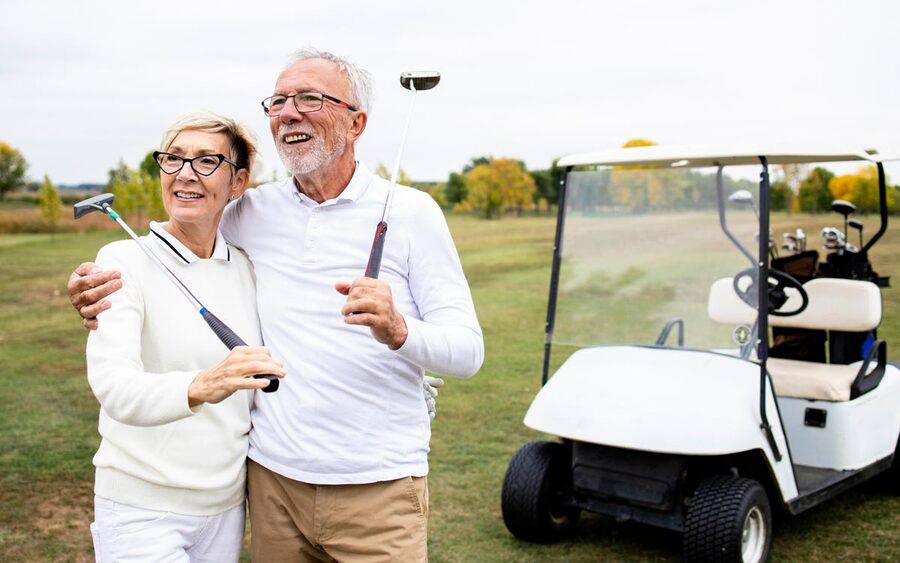 A mature couple holds golf clubs over their shoulders next to a golf cart.