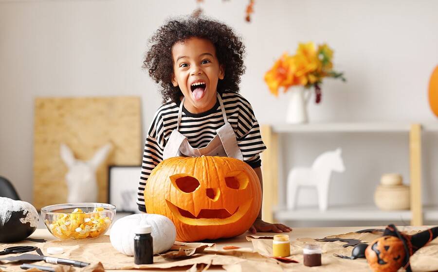 A young child makes a scary face standing over carved Halloween pumpkin.