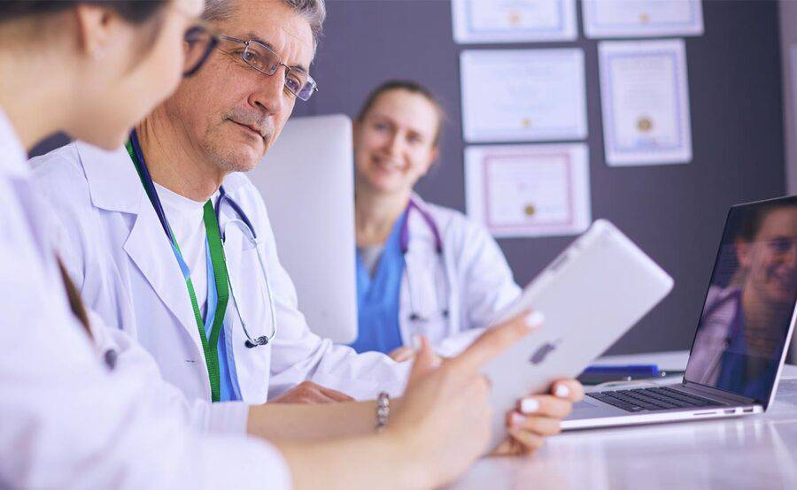 A group of physicians sits at a table with  tablets and laptops, representing medical education and training programs.