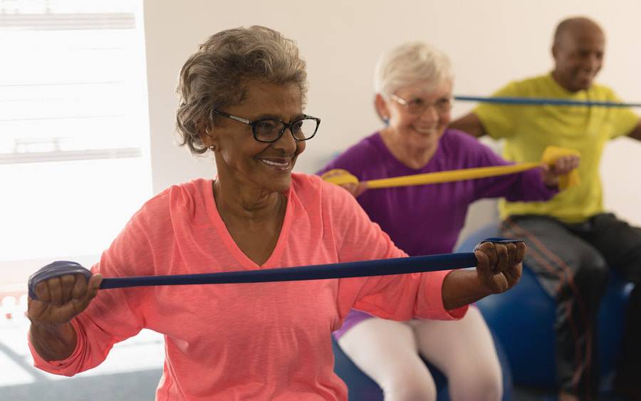 A diverse group of older men and women smile while participating in an exercise class using bands to improve their strength.