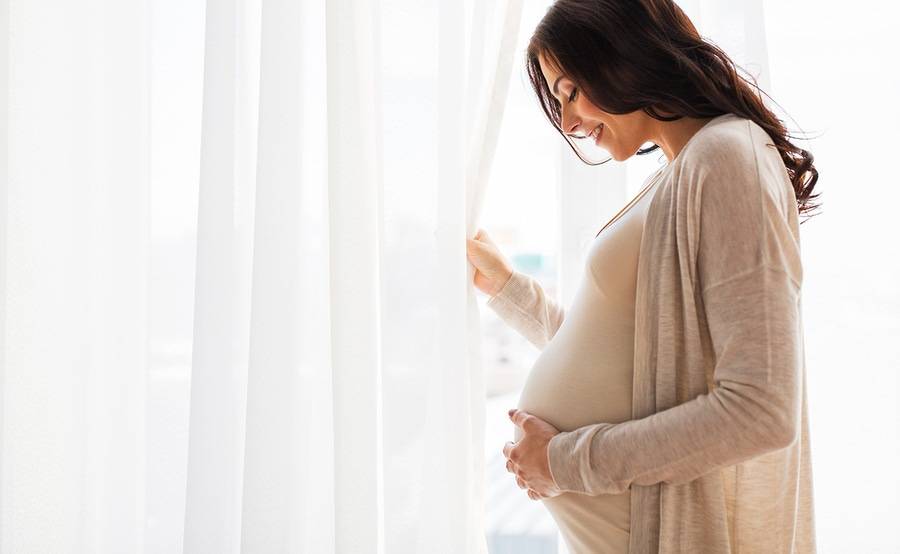A pregnant woman standing by a window in a sunlit room.