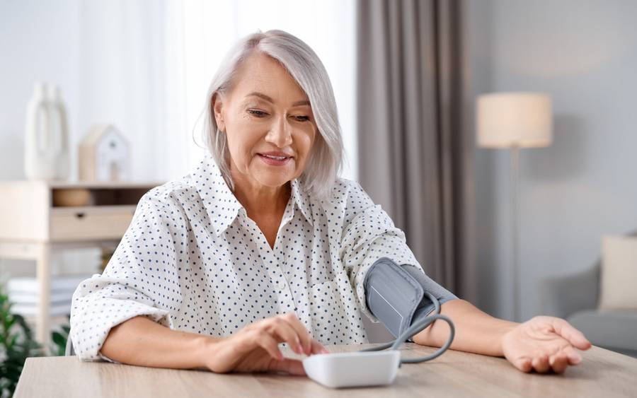 An older woman with short gray hair check her blood pressure while sitting at a table at home.