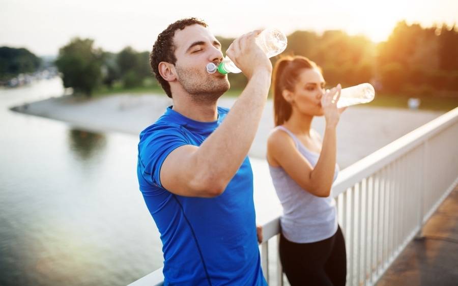 A fit couple in athleisure stand on a bridge overlooking a river at sunset and drink bottled water to stay cool and hydrated.
