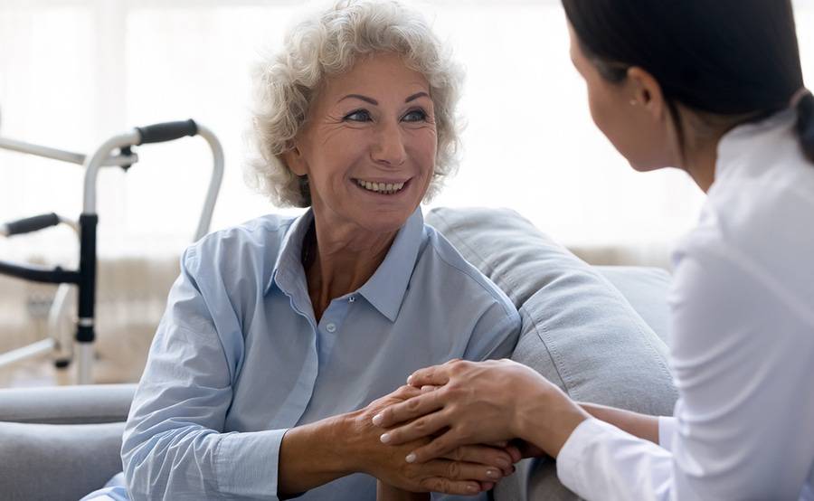 A senior woman on a sofa hold hands with a caregiver. A walker used for balance is in the background.