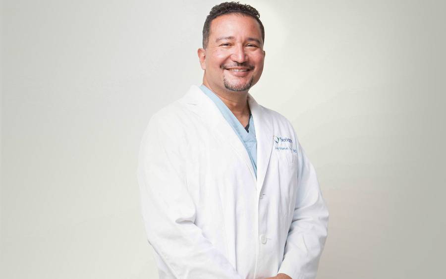 Juan Tovar, MD, a Hispanic/Latino male doctor, smiles in a white lab coat, proud of the SCHE's focus on health equity.