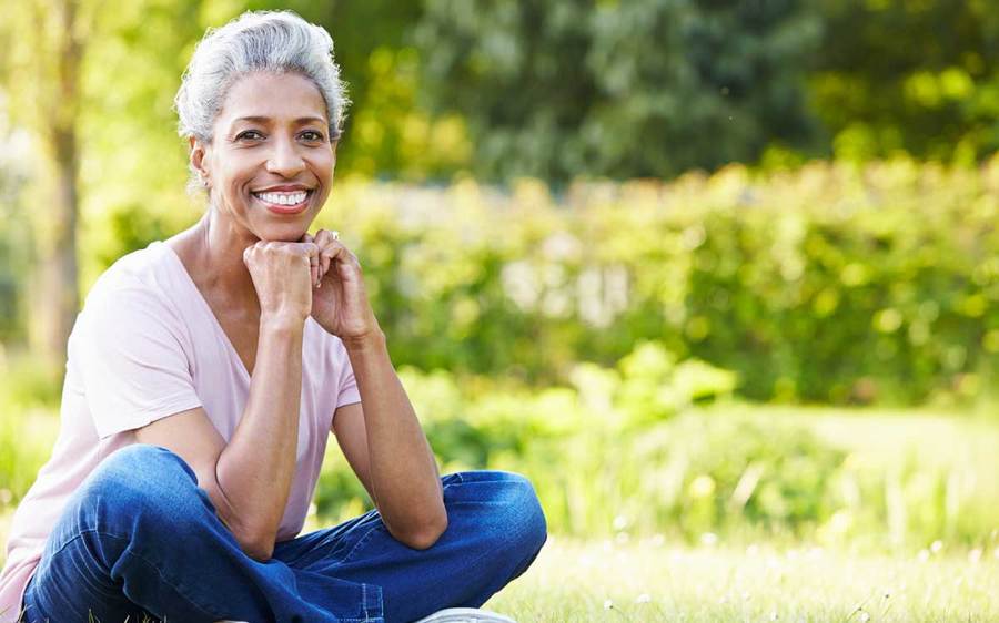 A smiling middle-aged woman wearing denim jeans sits cross-legged in a grassy park setting