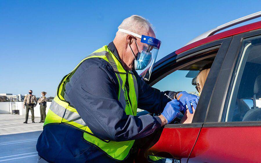 Scripps Health President and CEO Chris Van Gorder administers a COVID-19 vaccine to a law enforcement officer in their car.