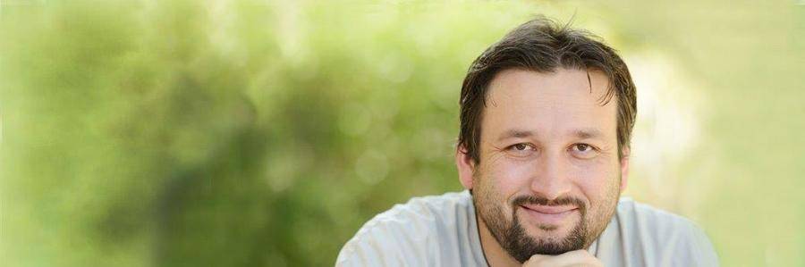 A bearded middle-aged man shares a small smile in a sunny garden setting.