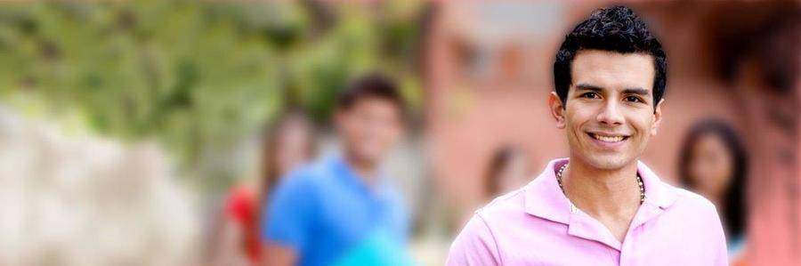 A happy young man pauses for a smile on a university walkway with fellow students and a college building visible in the background.