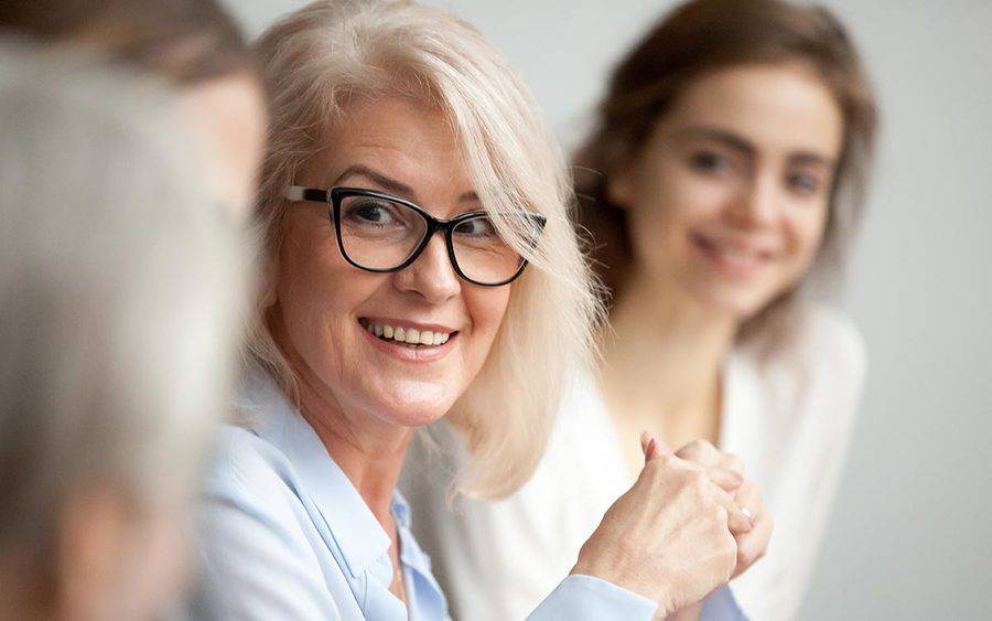 A smiling mature woman in glasses talks to a group of other adults, representing the importance of discussing lymphedema after cancer treatment.