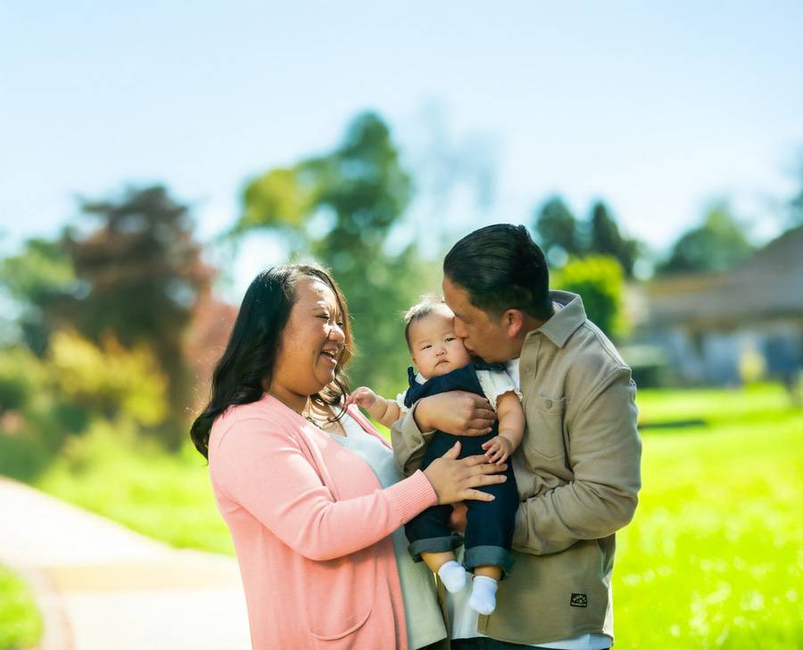 Mai Vue and husband Richard kiss their new baby girl Athena outdoors on a sunny day at a park.