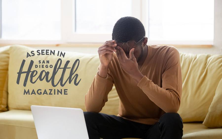 A man sitting on a couch with his latop across from him, rubbing his eyes, demonstrating his eyes are dry caused by over using digital devices. 