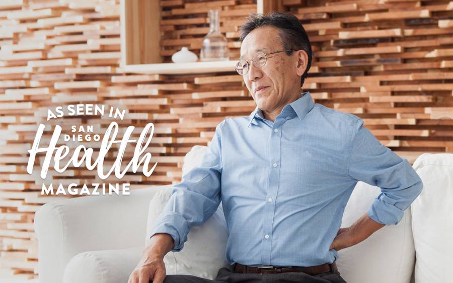 A mature man holds his back in pain while sitting on a couch, representing kidney stone symptoms.