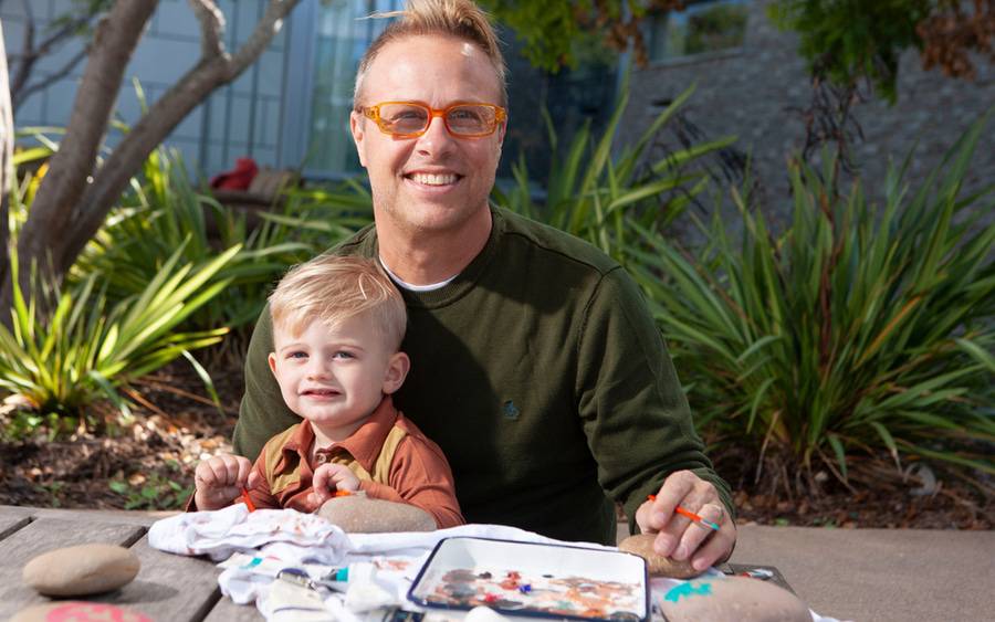 Painter and father, Mark DeSalvo, wears a green sweater with his toddler son in his lap, and is happy to beat tonsil cancer.