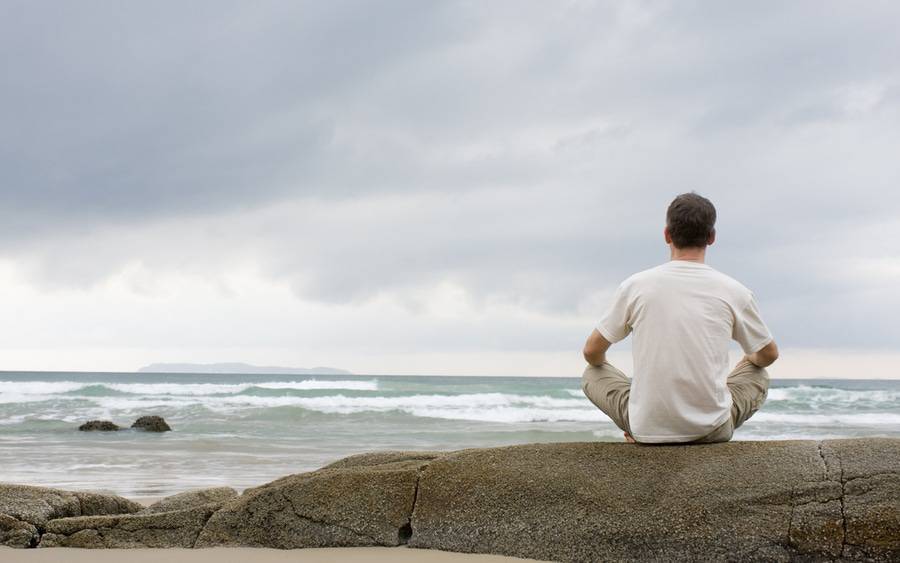 A man sits on a rock by the beach, practicing mindfulness-based stress reduction.