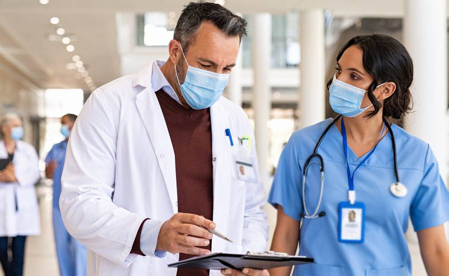 Two medical professionals in surgical masks talk while walking down a hospital hallway.