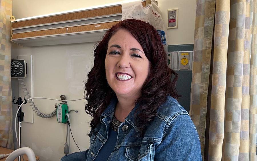 Melody Thomas smiles in a denim jacket in a hospital room, with medical equipment and patterned curtains behind her.