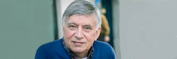 Man with blue eyes and gray hair wearing dark blue sweater and plaid collared shirt.