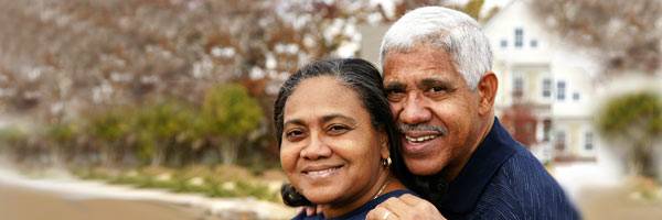 Gray haired man and dark haired woman wearing dark blue clothing and embracing with large white home and trees in the background.
