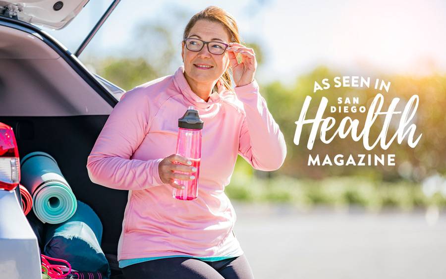 A woman sits in the back of her SUV, drinking water, with fitness gear in view including a jump rope and a yoga mat. SD Health Magazine