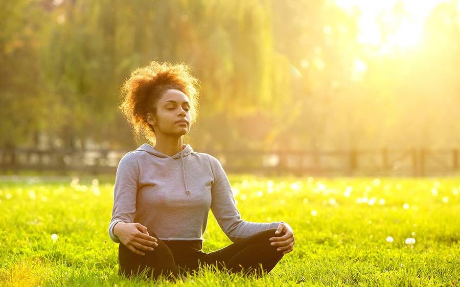 A young and fit African American woman mediates in a wide open field as a way to ease stress and avoid burnout.