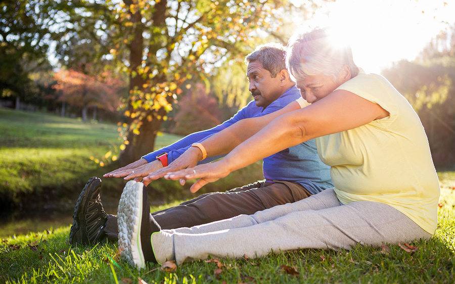 A healthy older couple reach for their toes and stretch out on the grass at park on a sunny day.