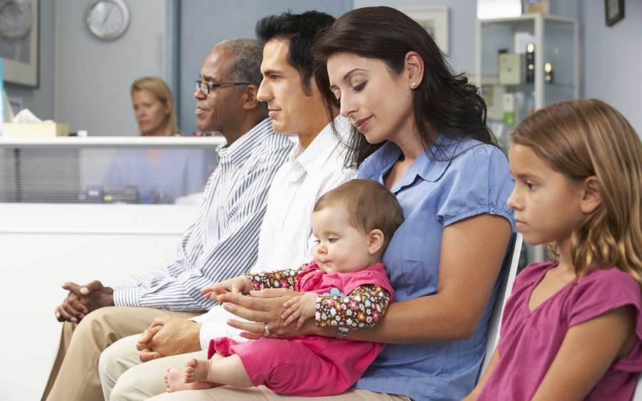 Three adults and two children sit in a doctor's waiting room.