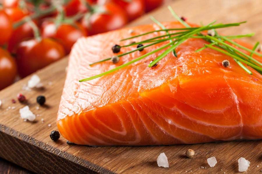 A fillet of salmon, one of the healthier kinds of fish to eat, lies on a cutting board sprinkled with fresh herbs and spices,