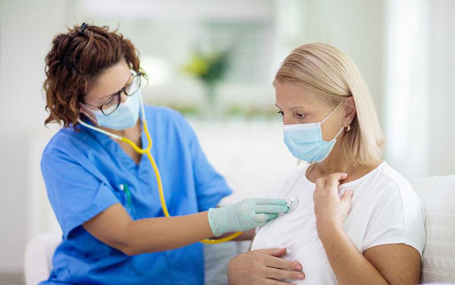 A nurse in mask and gloves checks a woman's heart rate while she holds her chest and neck, representing persistent COVID symptoms.