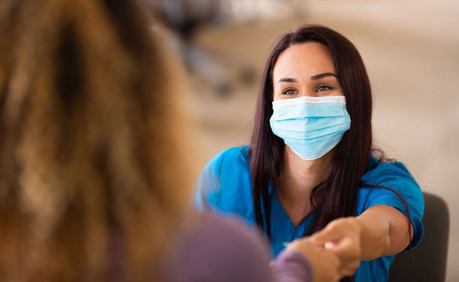 A nurse checking-in a patient at a desk with a face mask on.