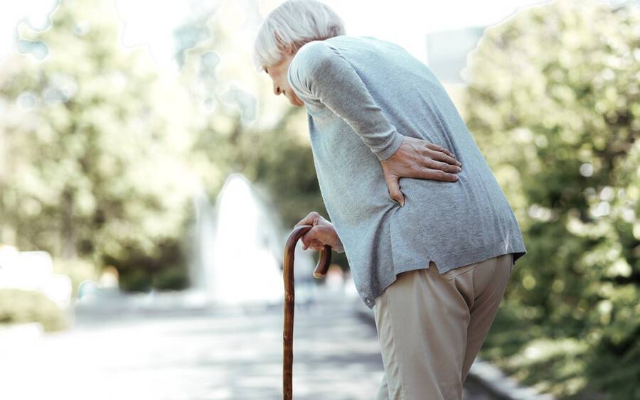 An older woman holding her back with one hand and grabbing walking cane with other.