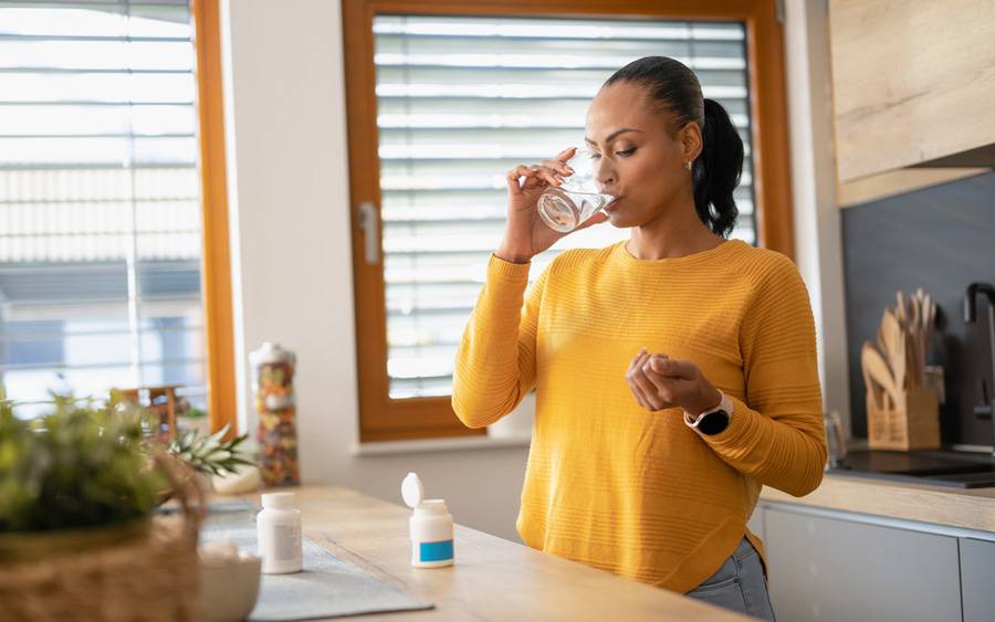 A woman stands in her kitchen drinking a cup of water as she swallows a pill to treat cancer.