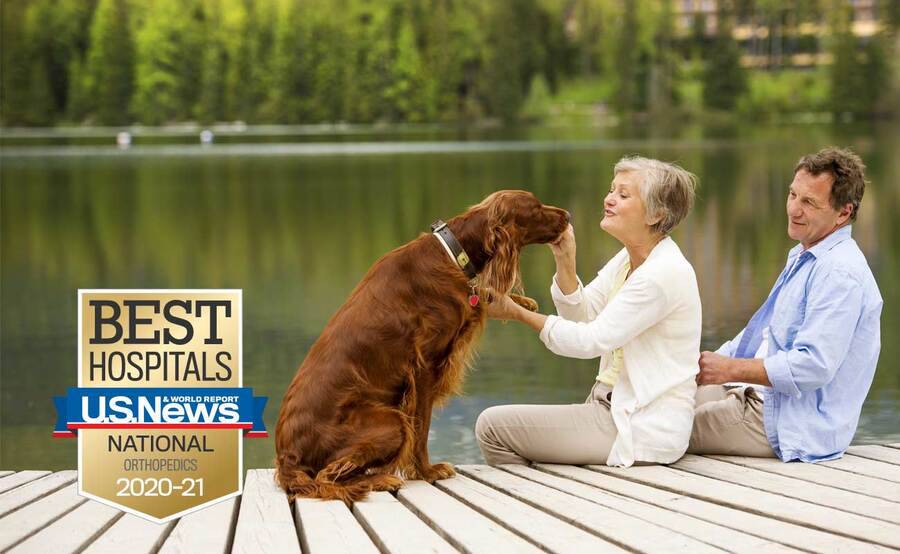 A mature woman and her partner sit on a dock with a golden retriever, representing an active lifestyle after joint reconstruction surgery.