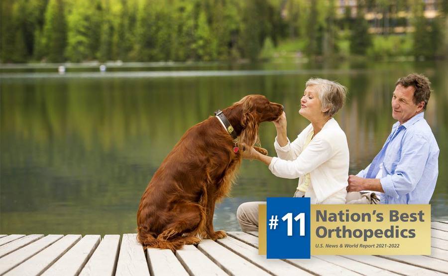 A mature woman and her partner sit on a dock with a golden retriever, representing an active lifestyle after joint reconstruction surgery.