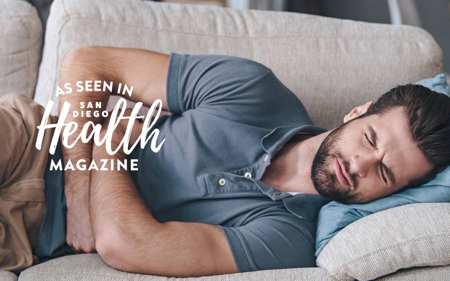 A young white man lies on the couch with his arms wrapped around his stomach and his eyes closed in pain.