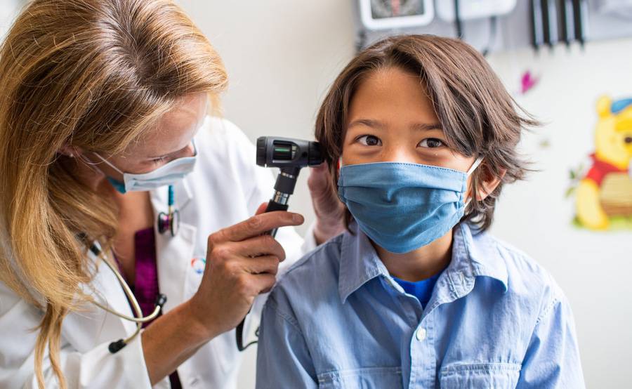 A school-aged child sits patiently while a doctor looks inside their ear with an otoscope.