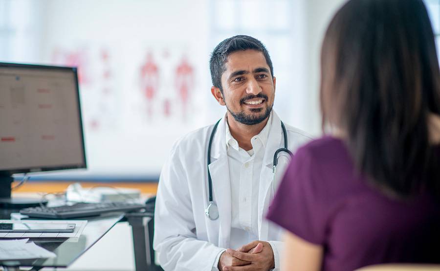 A physician speaking with a patient in an office with a computer and charts of the human body.