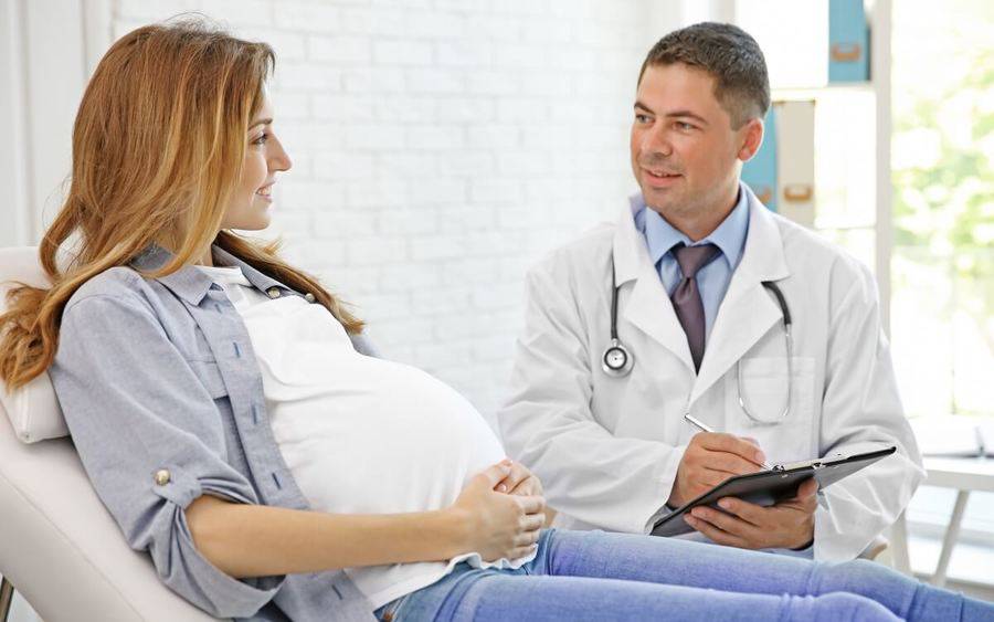 Gynecologist talks to young pregnant woman sitting on bed.