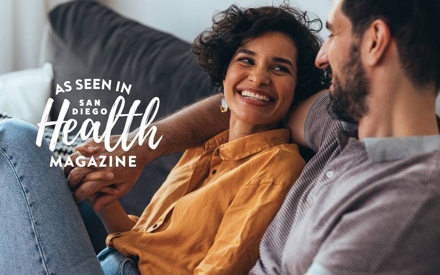 A man and a woman smile as they sit on the couch and hold hands - SD Health Magazine
