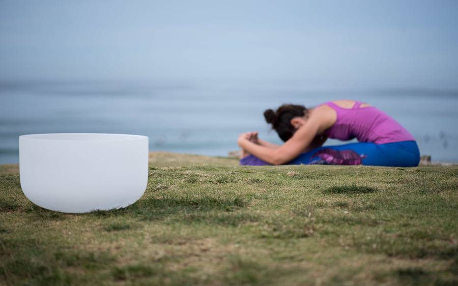 A woman practices a seated yoga pose in the background next to a singing bowl, representing the healing atmosphere of restorative yoga.