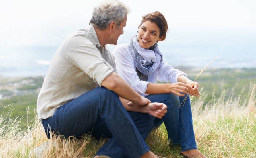 A woman and a man sitting on ground covered with tall grass atop a hill.