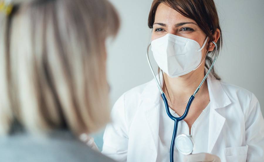 A doctor smiles behind a surgical mask while preparing to listen to a patient's heart with a stethoscope.