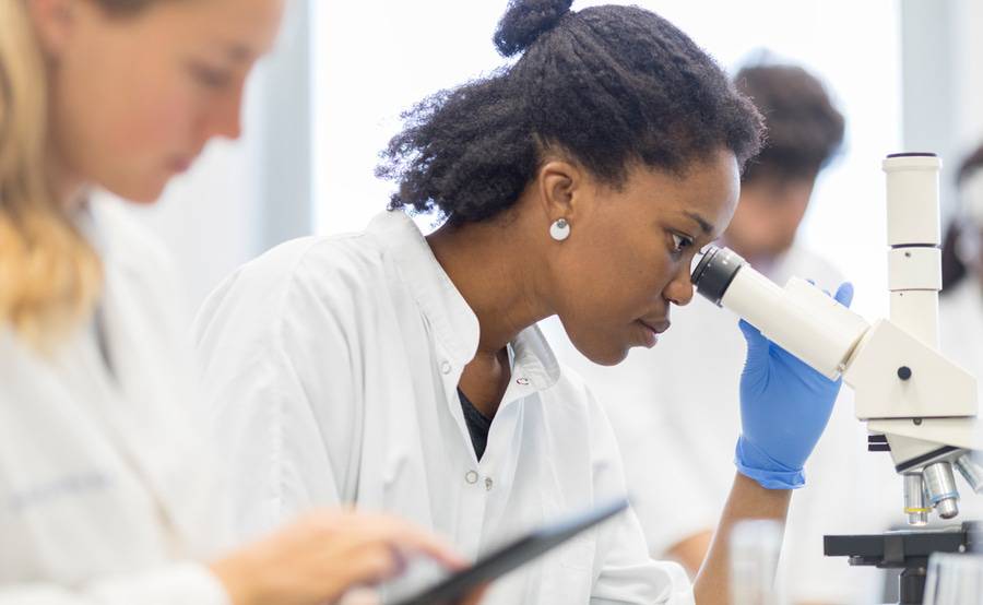 A black person looks through a microscope, representing revised research efforts on health care disparities.