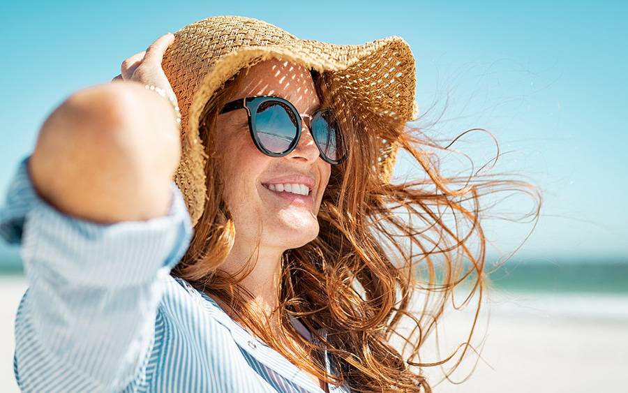 A red-haired woman in sunglasses and a wide brim hat enjoys the sun, representing skin safety tips from a Scripps expert.