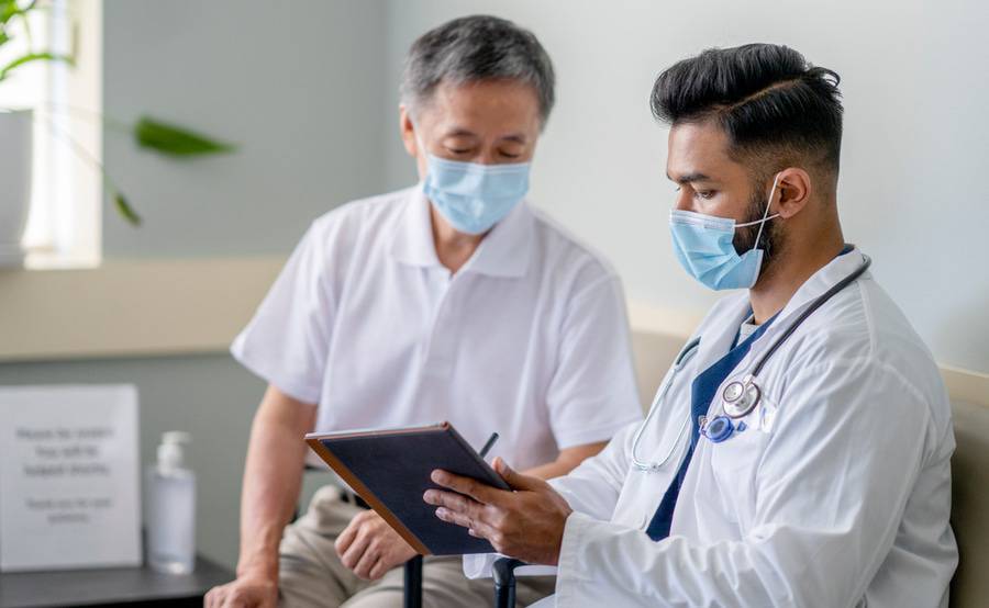 A physician in a surgical mask sits next to a patient in a waiting, explaining information from a clipboard.